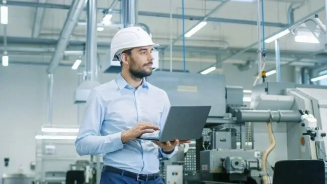 Engineer in a hard hat reviewing systems on a laptop in a manufacturing facility